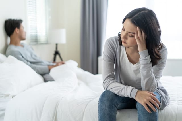 A woman sitting on the edge of a bed, looking down with a hand on her forehead in a gesture of sadness or distress, while another person sits in the background, suggesting a moment of emotional pain related to a relationship breakup.