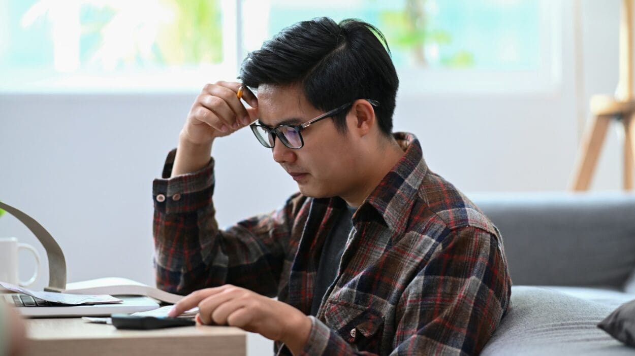 A man sitting at a table, looking troubled as he holds his glasses, symbolizing the burden of grief coupled with financial stress.