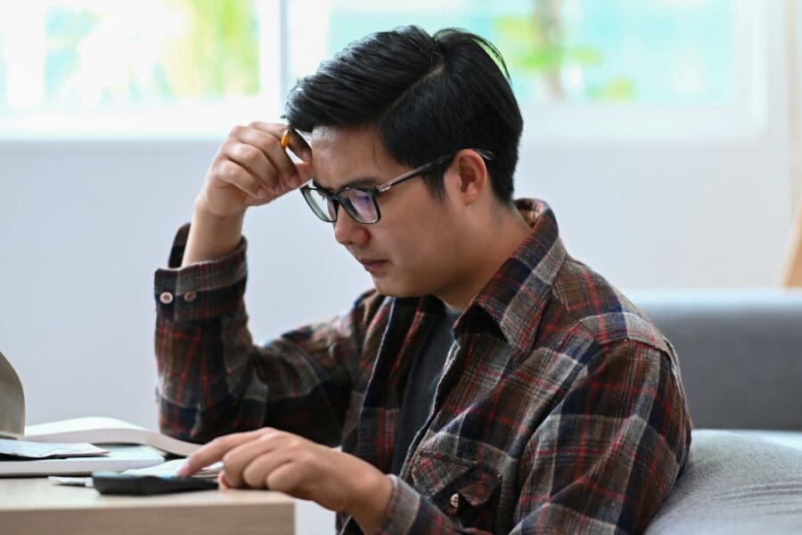 A man sitting at a table, looking troubled as he holds his glasses, symbolizing the burden of grief coupled with financial stress.