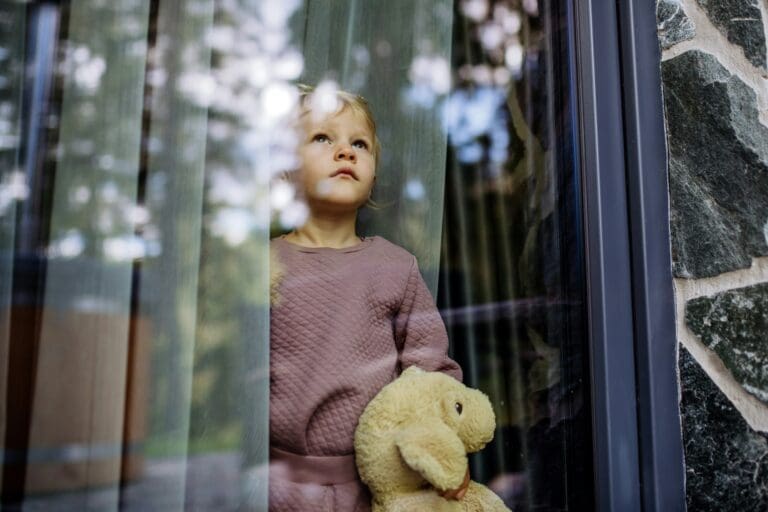 A young child holding a stuffed animal, gazing thoughtfully out a window, symbolizing the quiet, contemplative nature of children's grief and the need for gentle support and understanding.