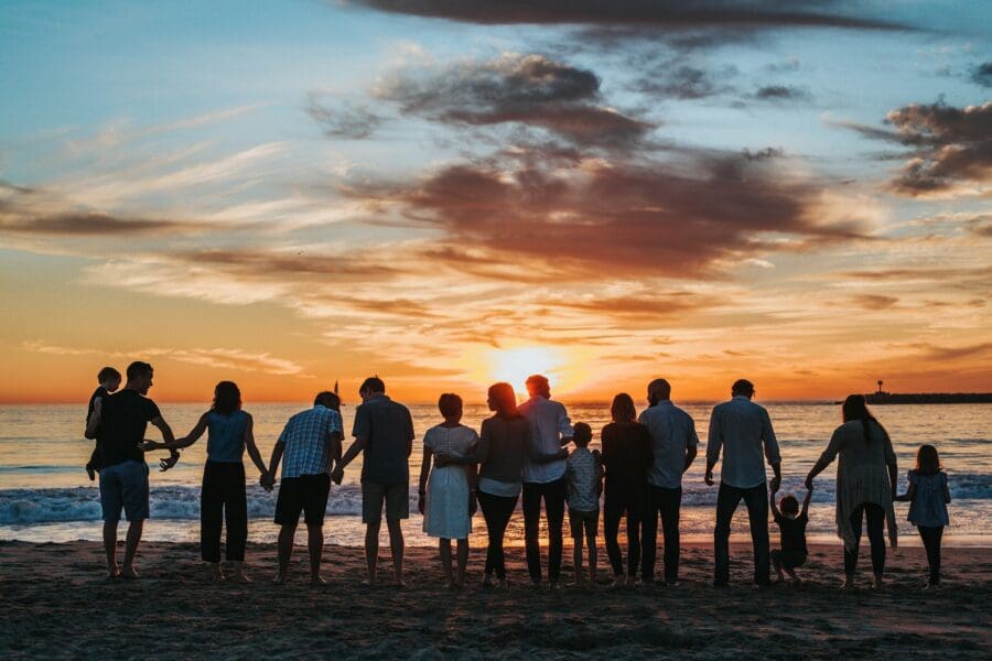 A large family holding hands and standing together on the beach at sunset, symbolizing the support and connection shared across generations during times of grief.