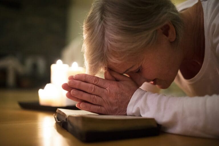 An elderly woman praying with her head bowed and hands clasped in front of an open book and lit candles, symbolizing the solace and strength found in faith during times of grief.