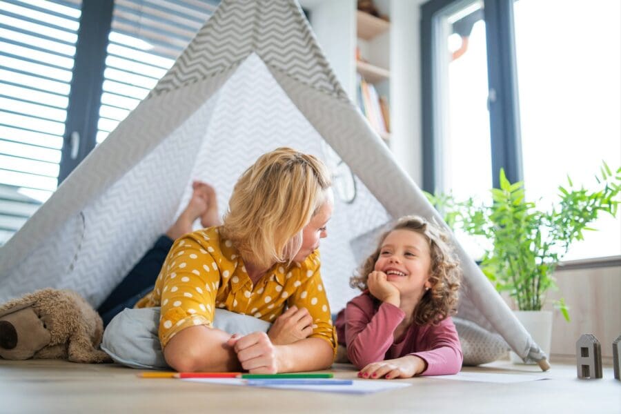 A single parent and child lying on the floor under a makeshift tent, smiling and engaging in a playful conversation. The scene is bright and cheerful, highlighting the bond and joy in their relationship amidst the challenges of single parenthood.