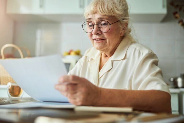 Elderly woman with glasses reviewing documents at her kitchen table, symbolizing vigilance against financial scams during times of grief.