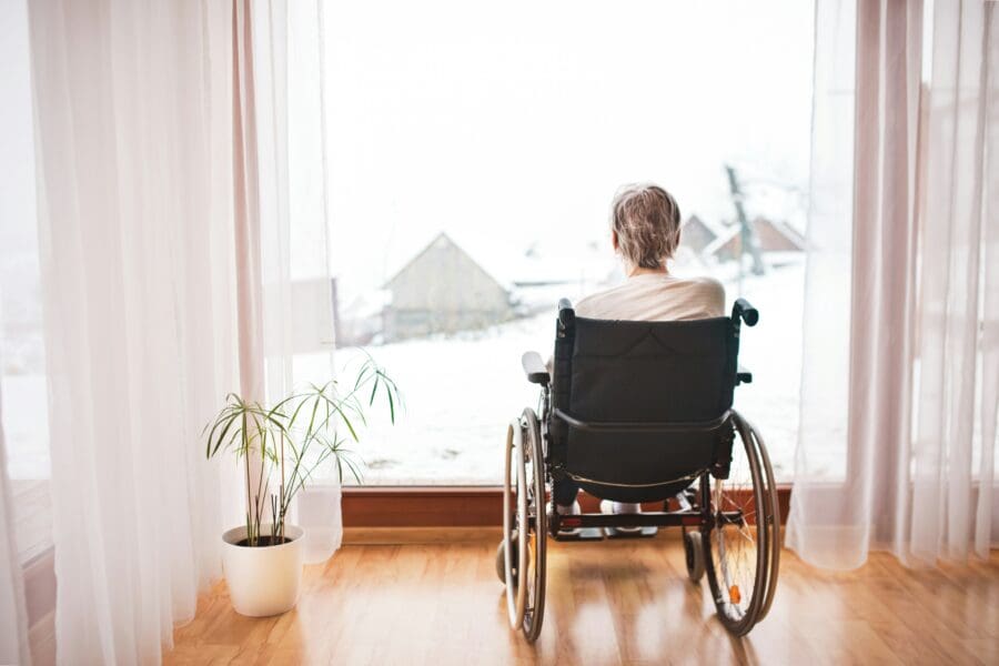 Senior person in a wheelchair looking out a window onto a snowy landscape, symbolizing the emotional distance and grief experienced by those with a parent living with dementia.