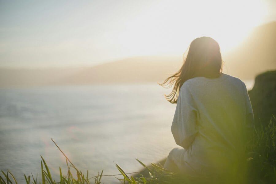 A woman sitting alone at the edge of a cliff, gazing out over the ocean at sunset, reflecting on the bittersweet passage of time and the enduring love behind the grief of losing her mother a year ago.