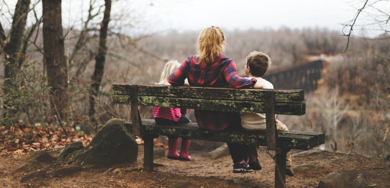 Mother sitting on a bench outdoors with two young children, looking out over a misty forest, symbolizing reflection and the emotional journey of grief and healing.