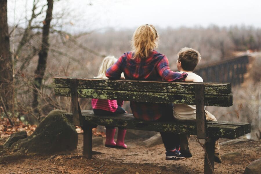 Mother sitting on a bench outdoors with two young children, looking out over a misty forest, symbolizing reflection and the emotional journey of grief and healing.