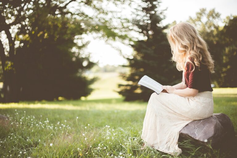 A woman sitting on a rock in a peaceful, sunlit meadow, deeply engrossed in reading a book about spirituality and the soul's journey after death.