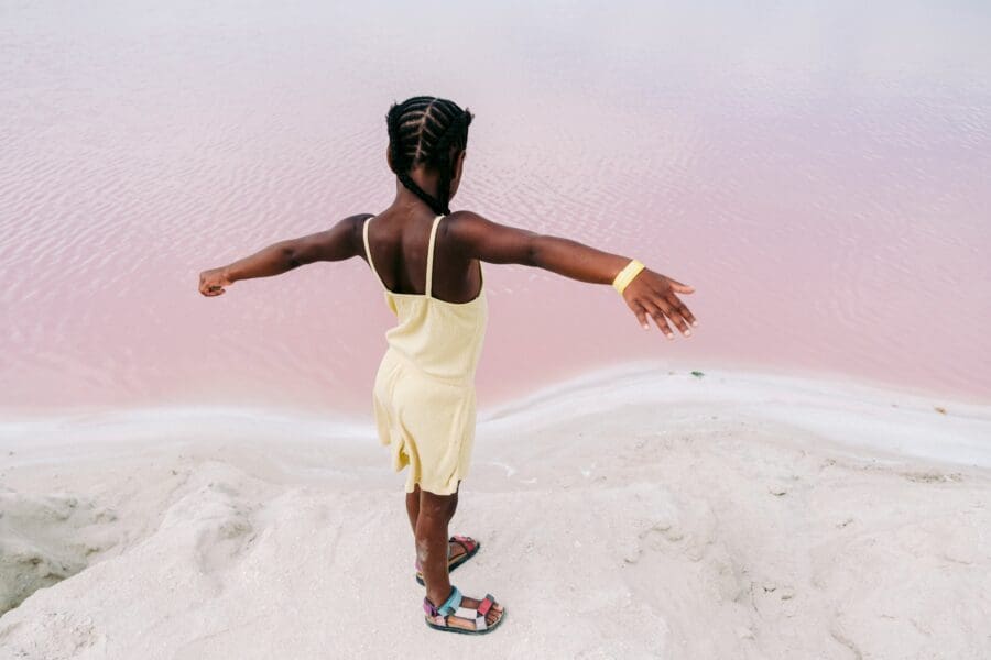 A young girl in a yellow dress joyfully extends her arms beside a pink-hued lake, symbolizing a moment of freedom and escape from a childhood home marked by emotional distance and parental discord.