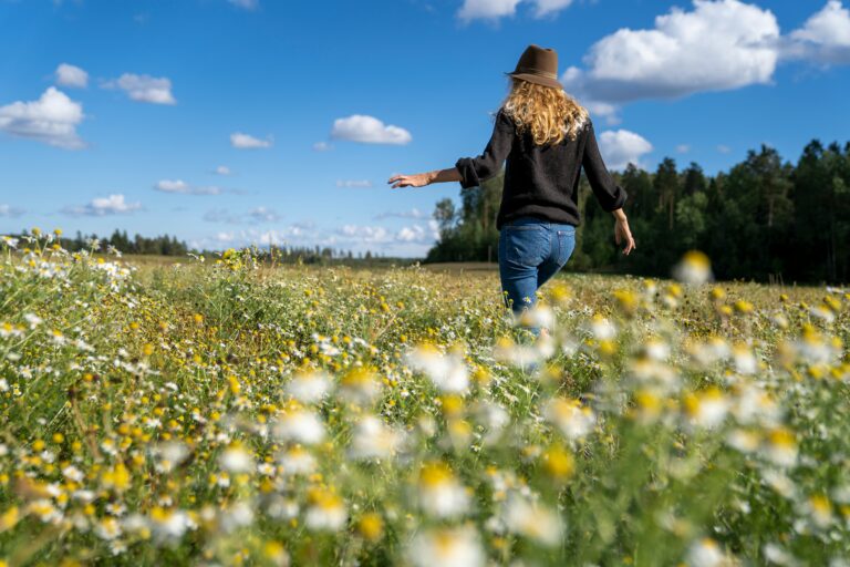 A woman in a black sweater and jeans, wearing a brown hat, walks through a lush meadow filled with white and yellow wildflowers, symbolizing renewal and moving forward in life.