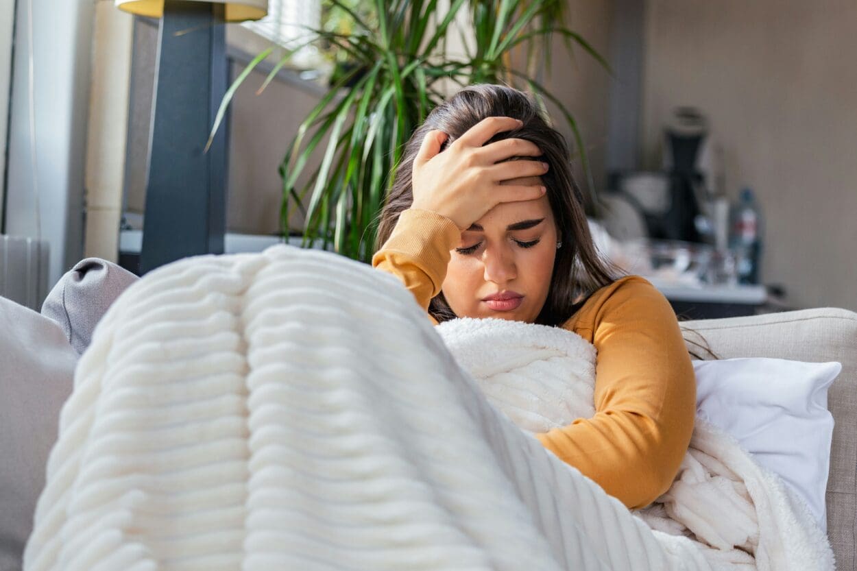 A distressed young woman sitting on a couch, clutching her head in pain, illustrating the physical manifestations of grief such as headaches and exhaustion.