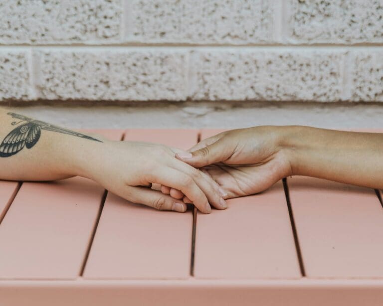 Two hands gently holding each other over a pink table, symbolizing the comfort and support shared between individuals coping with the profound psychological impact of losing a loved one to suicide.