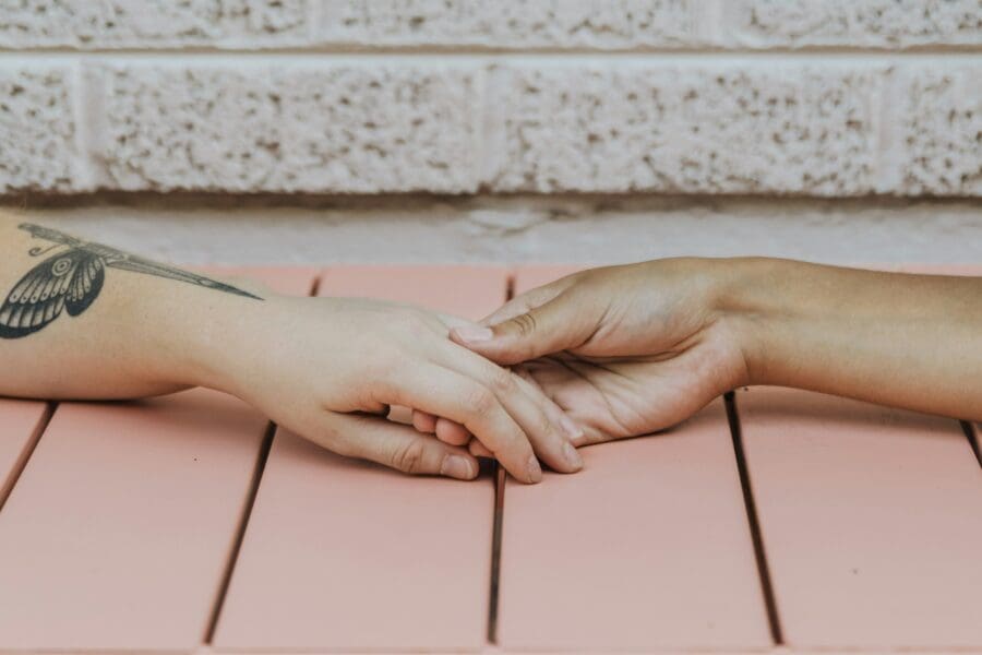 Two hands gently holding each other over a pink table, symbolizing the comfort and support shared between individuals coping with the profound psychological impact of losing a loved one to suicide.