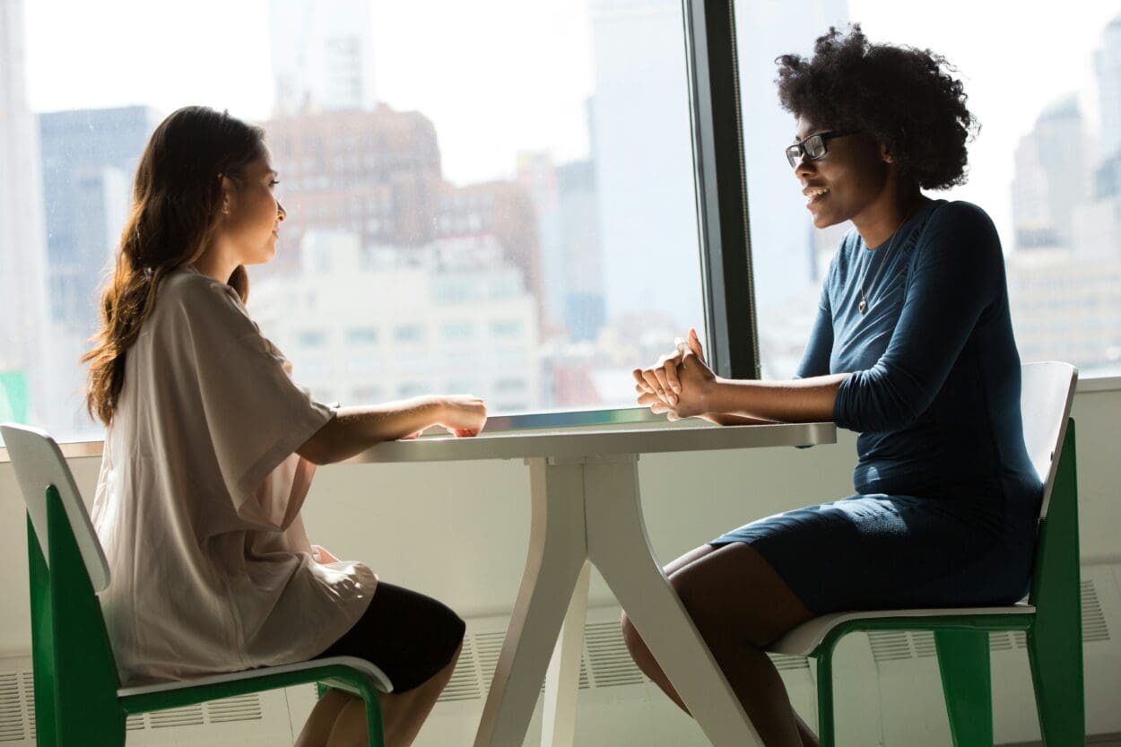 Two women engaging in a serious conversation at a table by a window overlooking the city, highlighting the crucial importance of discussing suicide and mental health openly to promote understanding and prevention.