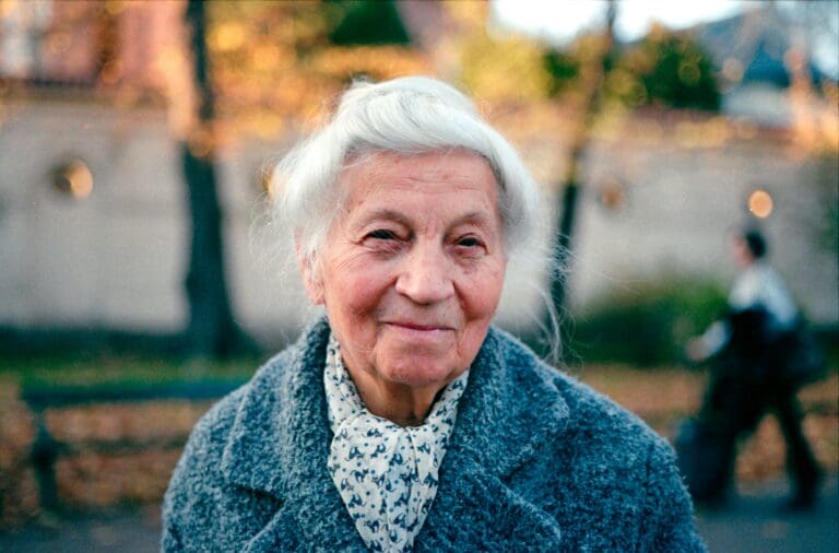 Portrait of an elderly woman with white hair and a gentle smile, wearing a cozy grey sweater and a patterned scarf, standing outdoors with autumn leaves in the background. This image represents the heartache of moving abroad and leaving behind a loved one with dementia.