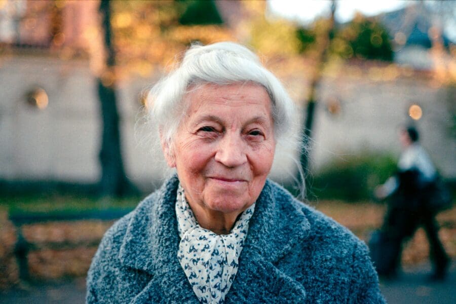 Portrait of an elderly woman with white hair and a gentle smile, wearing a cozy grey sweater and a patterned scarf, standing outdoors with autumn leaves in the background. This image represents the heartache of moving abroad and leaving behind a loved one with dementia.